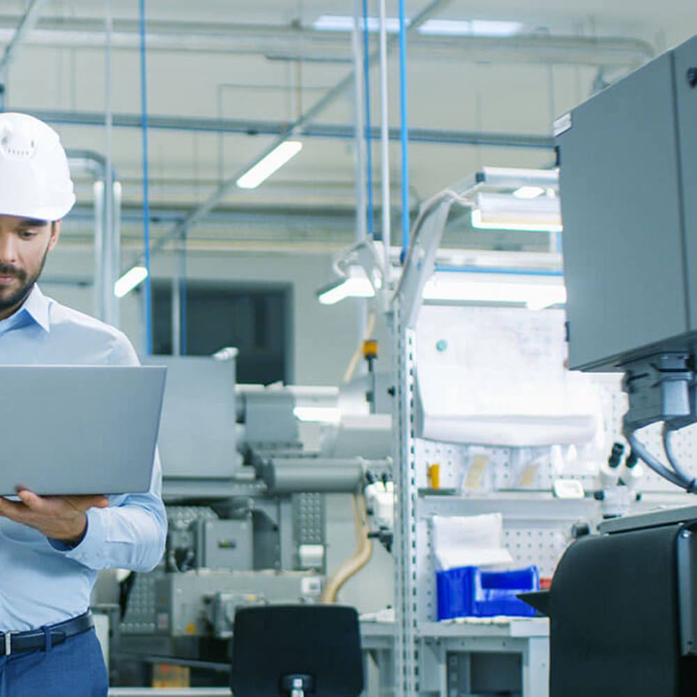 Chief Engineer in the Hard Hat Walks Through Light Modern Factory While Holding Laptop. Successful, Handsome Man in Modern Industrial Environment.