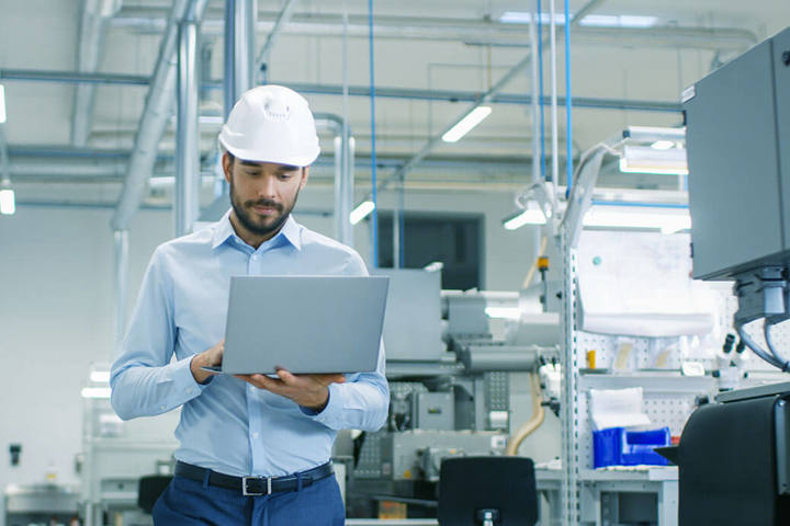 Chief Engineer in the Hard Hat Walks Through Light Modern Factory While Holding Laptop. Successful, Handsome Man in Modern Industrial Environment.
